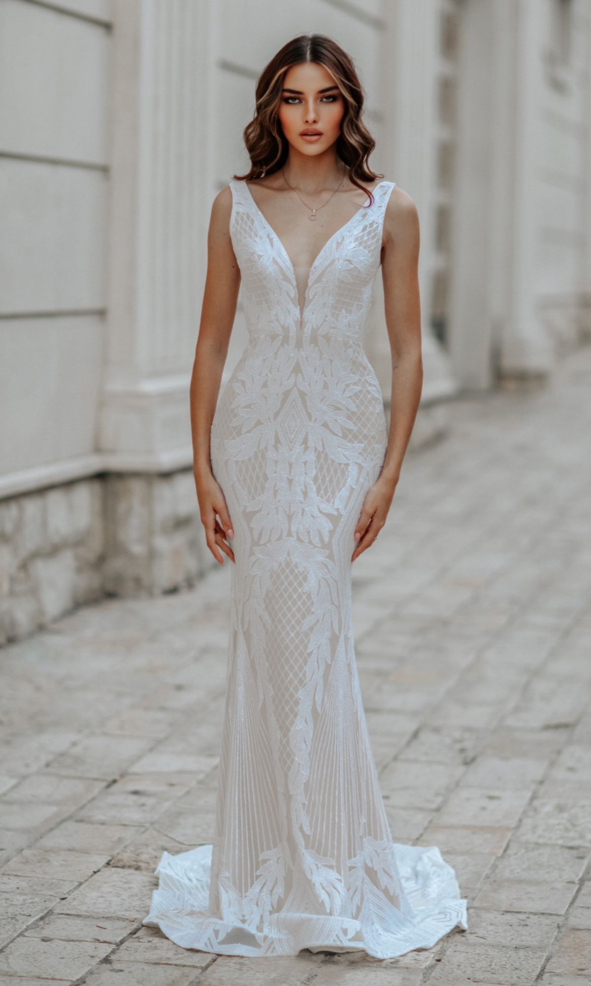 A woman models the Classic Long White Formal Dress: Tina Holly BB052, featuring a fitted silhouette, deep V-neckline, and intricate lace patterns, standing on a stone walkway by a light-colored building.